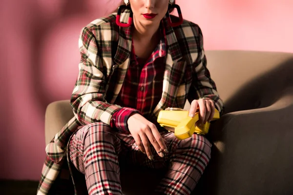 Cropped view of elegant girl in checkered suit posing in armchair with retro telephone on pink background — Stock Photo