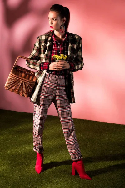 Hermosa chica elegante en traje a cuadros de otoño posando con cesta y flores en rosa - foto de stock