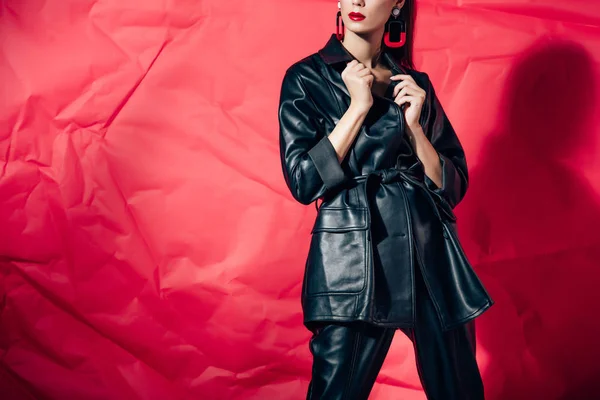 Обрезанный вид стильной женщины, позирующей в черном кожаном костюме на красном фоне — стоковое фото