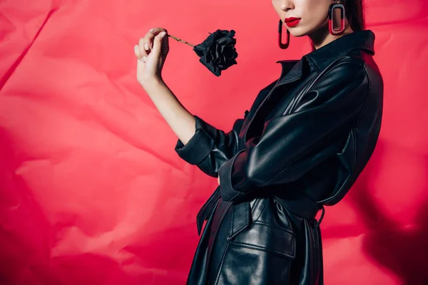 Vista recortada del modelo en traje de cuero negro posando con rosa negra sobre fondo rojo - foto de stock