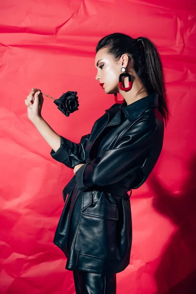 Beautiful young woman in black leather suit posing with black rose on red background — Stock Photo