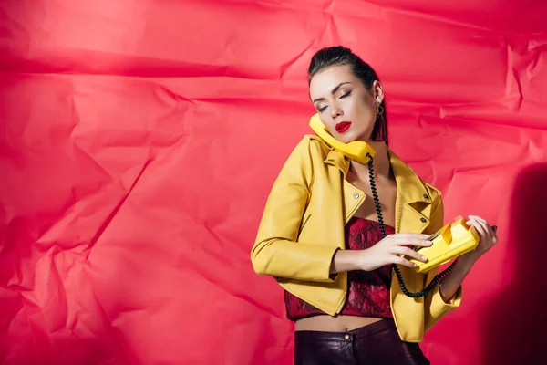 Hermosa mujer en chaqueta de cuero posando con teléfono rotatorio amarillo sobre fondo rojo - foto de stock