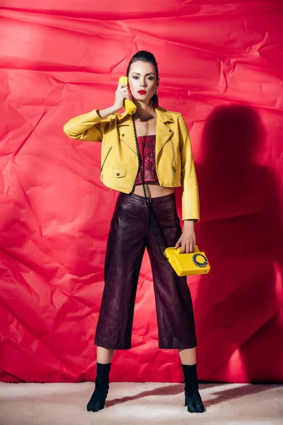 Young woman in yellow leather jacket talking on retro phone on red background — Stock Photo