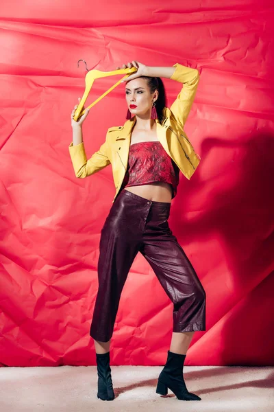 Stylish young woman in yellow leather jacket posing with hanger on red background — Stock Photo