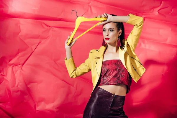 Beautiful stylish model in yellow leather jacket posing with hanger on red background — Stock Photo