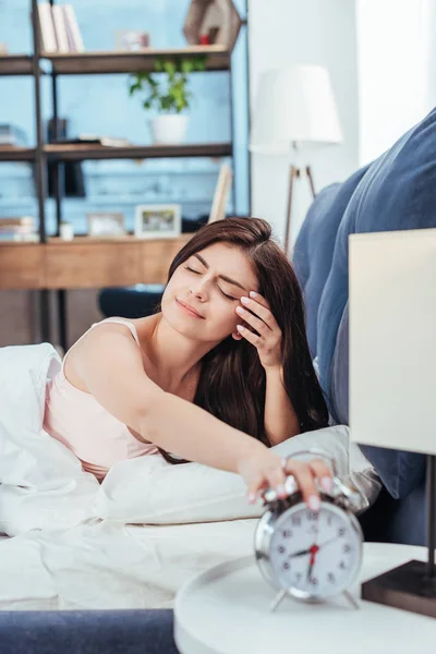 Сонная девушка выключает будильник, лежа на кровати в утреннее время дома — стоковое фото
