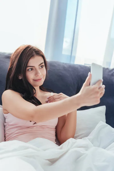 Щаслива жінка бере селфі на смартфон, сидячи на ліжку в ранковий час вдома — Stock Photo