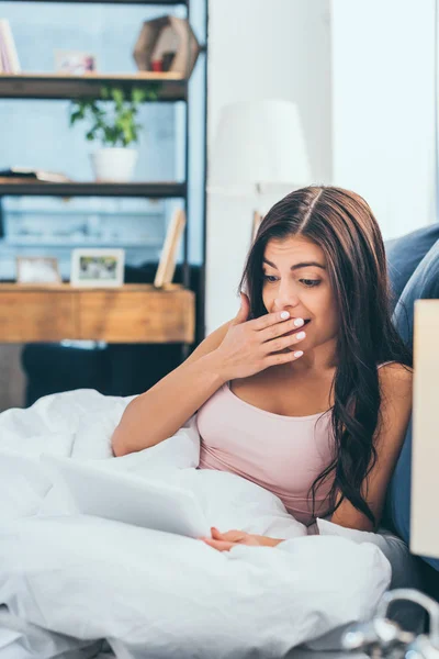 Überraschte junge Frau, die den Mund mit der Hand bedeckt und morgens zu Hause ein digitales Tablet im Bett benutzt — Stockfoto