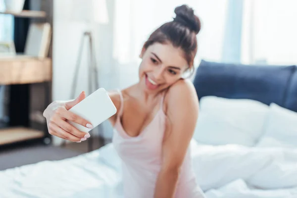 Chica feliz tomando selfie en el teléfono inteligente mientras está sentado en la cama durante la mañana en casa - foto de stock