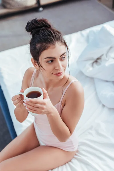 Високий кут зору щасливої молодої жінки, що сидить з чашкою кави на ліжку в ранковий час вдома — стокове фото