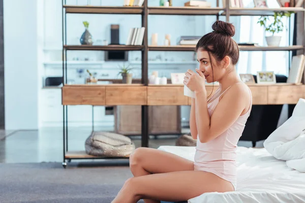 Вид сбоку на молодую женщину, сидящую с чашкой кофе на кровати в утреннее время дома — стоковое фото