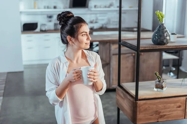 Menina sorridente em camisa branca segurando xícara de café durante a manhã em casa — Fotografia de Stock