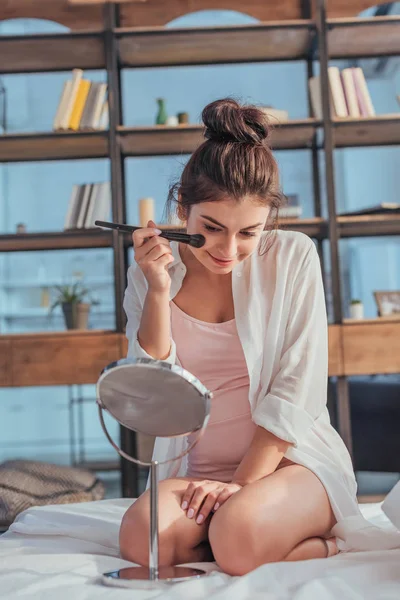 Schönes Mädchen, das zu Hause Spiegel anschaut und sich mit dem Pinsel auf dem Bett schminkt — Stockfoto