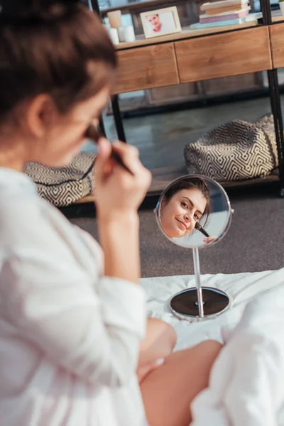Foco seletivo da menina bonita aplicando maquiagem por escova e olhando espelho na cama em casa — Fotografia de Stock