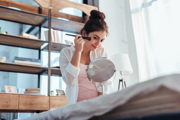 Low angle view of pretty girl applying makeup by brush and looking at mirror on bed at home — Stock Photo