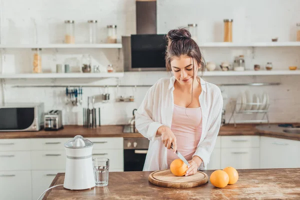 Pretty young woman cutting orange by knife on wooden board in kitchen — Stock Photo