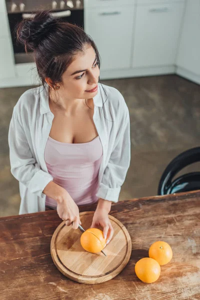 High angle view of attractive girl cutting orange by knife on wooden board in kitchen — Stock Photo