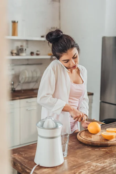 Mujer bonita cortando naranja por cuchillo y hablando en el teléfono inteligente en la cocina en casa - foto de stock