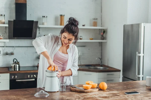 Chica sonriente haciendo jugo de naranja fresco con exprimidor en la mesa de madera en la cocina — Stock Photo