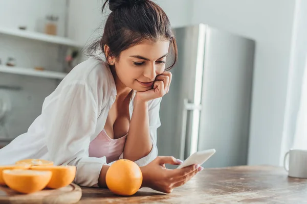 Selective focus of young woman using smartphone at wooden table with oranges in kitchen at home — Stock Photo