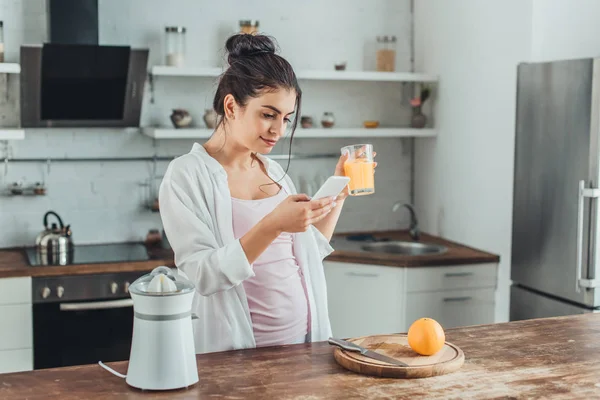 Attractive young woman using smartphone and holding glass with orange juice in kitchen at home — Stock Photo