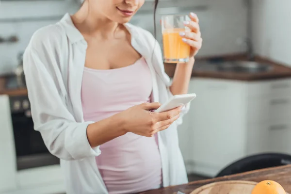 Частичный вид молодой женщины с помощью смартфона и проведение стекла с апельсиновым соком на кухне дома — стоковое фото
