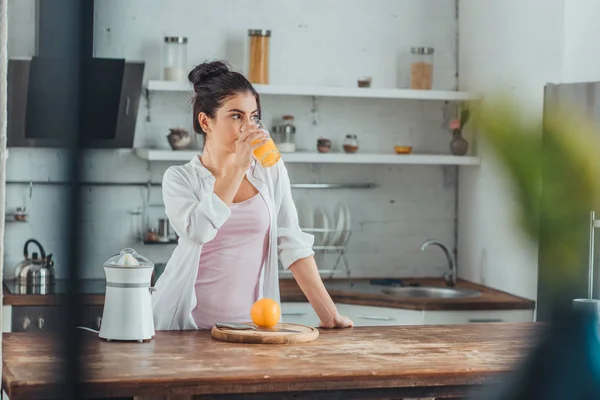 Mujer joven bebiendo jugo de naranja en la cocina durante la mañana en casa - foto de stock