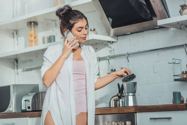 Jolie fille parler sur smartphone et vérifier cafetière dans la cuisine à la maison — Photo de stock