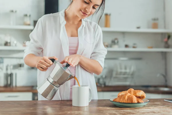 Красивая девушка наливает кофе в чашку за деревянным столом с круассанами на кухне дома — стоковое фото