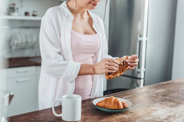 Partial view of woman having breakfast with croissants and coffee at wooden table in kitchen at home — Stock Photo