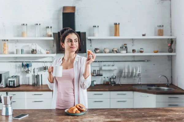 Jeune femme petit déjeuner avec croissants et café à table en bois dans la cuisine à la maison — Photo de stock