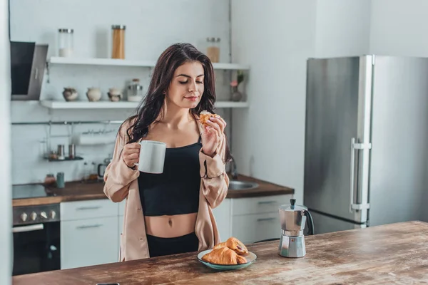 Chica bonita desayunando con croissants y café en la mesa de madera en la cocina en casa - foto de stock