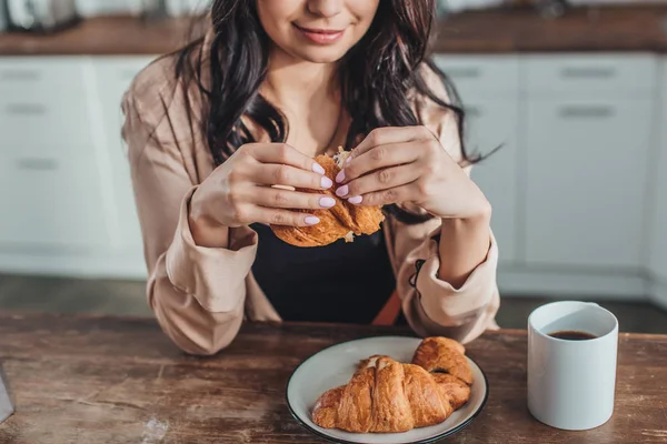 Foto ritagliata di ragazza che fa colazione con croissant e caffè al tavolo di legno in cucina a casa — Foto stock