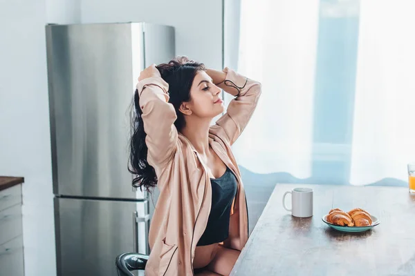 Atraente jovem mulher sentada à mesa de madeira com xícara de café e croissants na cozinha em casa — Fotografia de Stock