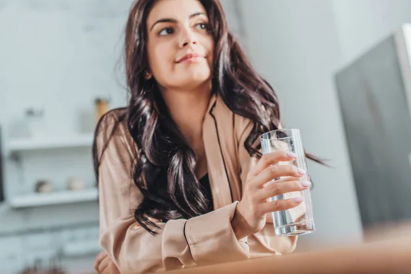 Low angle view of beautiful young woman holding glass with water and sitting at wooden tabletop in kitchen at home — Stock Photo