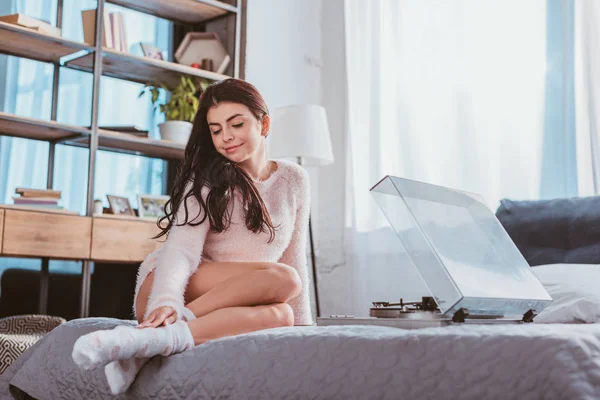 Привлекательная улыбающаяся девушка сидит на кровати и слушает виниловый аудиоплеер дома — стоковое фото