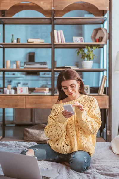 Joven freelancer en gafas usando smartphone mientras está sentada en la cama con laptop en casa - foto de stock