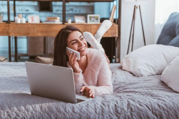 Rindo jovem freelancer feminino falando no smartphone enquanto deitado na cama com laptop em casa — Fotografia de Stock