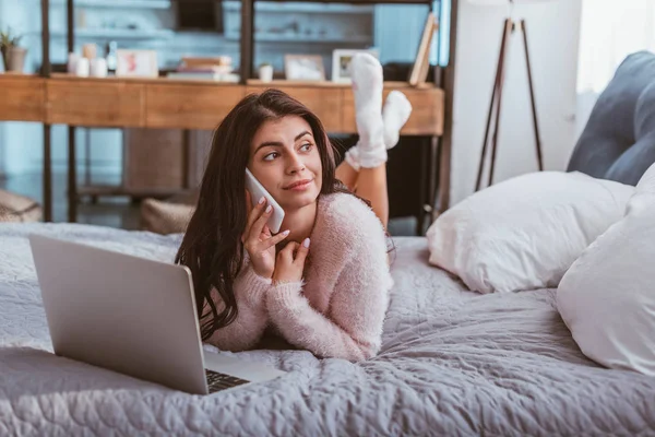 Atractiva joven freelancer hablando en el teléfono inteligente mientras está acostado en la cama con el ordenador portátil en casa - foto de stock