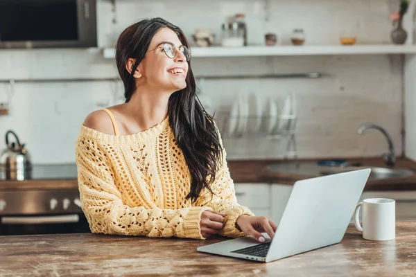 Laughing female freelancer in eyeglasses sitting at wooden table with laptop and coffee cup in kitchen at home — Stock Photo