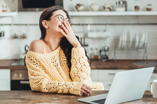 Selective focus of laughing female freelancer sitting at wooden table with laptop and coffee cup in kitchen at home — Stock Photo