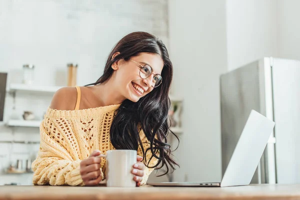Young laughing female freelancer sitting at wooden table with laptop and coffee cup in kitchen at home — Stock Photo
