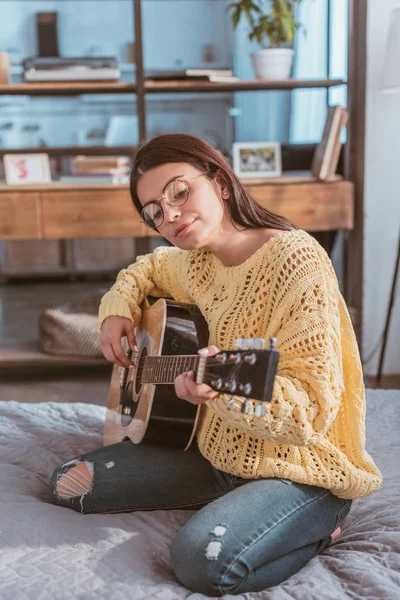 Mulher atraente em óculos tocando na guitarra acústica enquanto sentado na cama em casa — Fotografia de Stock