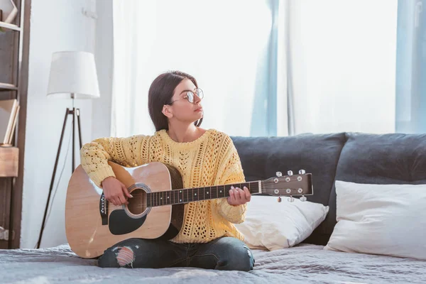 Jovem mulher bonita em óculos tocando na guitarra acústica enquanto sentado na cama em casa — Fotografia de Stock