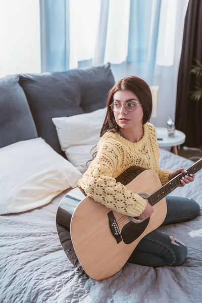 Vue grand angle de fille dans des lunettes de vue jouant sur la guitare acoustique tout en étant assis sur le lit à la maison — Photo de stock