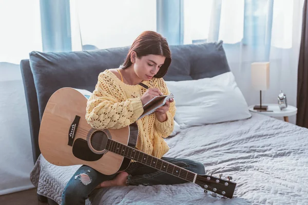 Menina bonita sentada com guitarra acústica e canção de escrita no livro didático na cama em casa — Fotografia de Stock