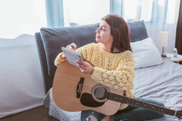 Happy girl sitting with acoustic guitar and textbook on bed at home — Stock Photo