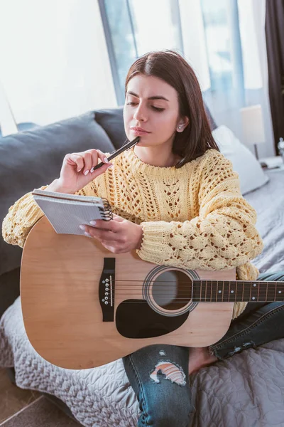 Niña pensativa sentada con guitarra acústica y libro de texto en la cama en casa - foto de stock