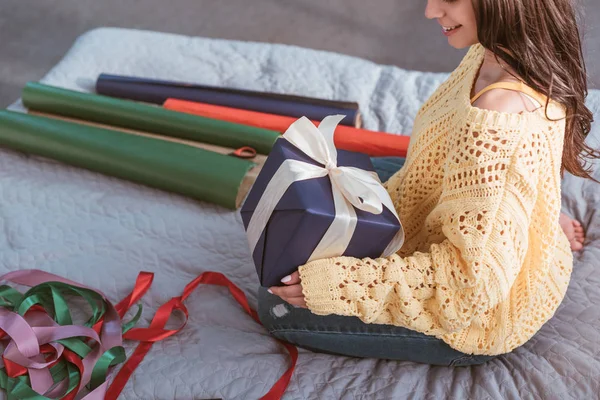 Vista parcial de la mujer joven sosteniendo caja de regalo envuelta en cinta mientras está sentada en la cama en casa - foto de stock