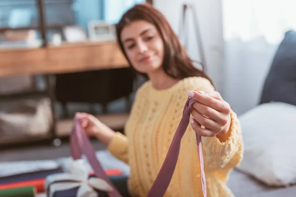 Selective focus of young woman holding festive ribbon while sitting on bed at home — Stock Photo